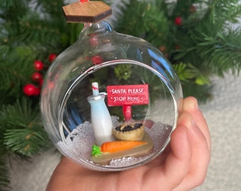 May include: A glass Christmas ornament with a miniature scene inside. The scene includes a bottle of milk, a carrot, a mince pie, and a sign that reads "Santa Please Stop Here".