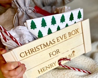 May include: A white wooden box with the text "Christmas Eve Box" and "For Henry" engraved on it. The box is filled with a white bag, tissue paper, candy canes, and a white napkin with green Christmas trees printed on it.