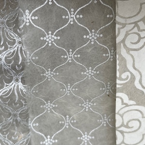 Könnte beinhalten: Drei Stücke Dekorpapier mit verschiedenen silbernen Mustern auf grauem Hintergrund. Die Muster umfassen ein florales Design, ein geometrisches Design und ein wirbelndes Design.
