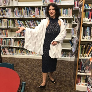 May include: A woman wearing a navy blue, patterned dress and a white cable-knit cardigan. She is standing in a library with bookshelves filled with books. She is wearing black shoes.