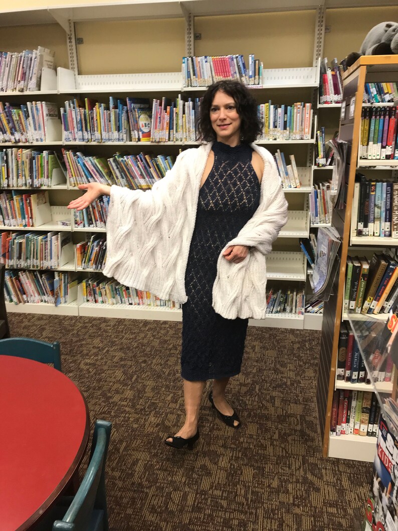 May include: A woman wearing a navy blue, patterned dress and a white cable-knit cardigan. She is standing in a library with bookshelves filled with books. She is wearing black shoes.