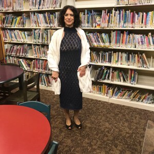 May include: A woman wearing a navy blue, open-weave dress with a high neckline and a white, cable-knit shawl. She is standing in a library with bookshelves filled with books. She is wearing black open-toe shoes.