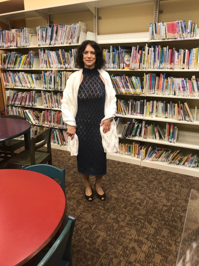 May include: A woman wearing a navy blue, open-weave dress with a high neckline and a white, cable-knit shawl. She is standing in a library with bookshelves filled with books. She is wearing black open-toe shoes.
