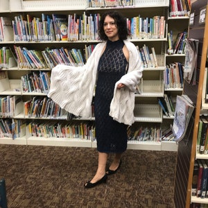 May include: A woman wearing a navy blue, sleeveless, knee-length dress with a diamond pattern, paired with a white, cable-knit shawl. She is wearing black open-toe shoes. The background is a library with bookshelves filled with books.