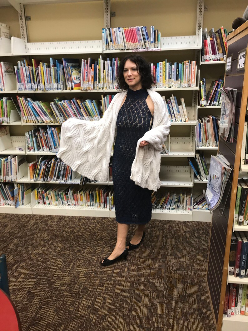 May include: A woman wearing a navy blue, sleeveless, knee-length dress with a diamond pattern, paired with a white, cable-knit shawl. She is wearing black open-toe shoes. The background is a library with bookshelves filled with books.