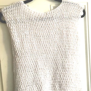 May include: A sleeveless top crafted from white yarn in an open, diamond-shaped crochet knit pattern. The garment features a wide, rounded neckline and a straight hem, showcasing a lightweight, semi-sheer construction with a consistent texture.