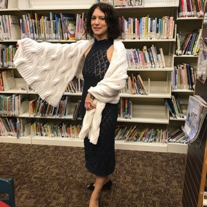 May include: A woman wearing a navy blue, open-weave dress and a white cable-knit shawl. The dress has a high neckline and a fitted silhouette. The shawl is draped over the shoulders and arms. Black open-toe heels complete the outfit. The background is a library.