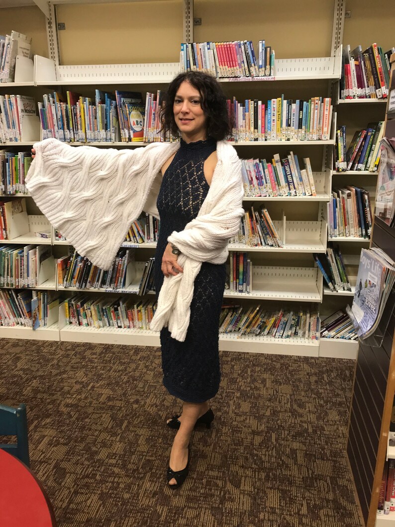 May include: A woman wearing a navy blue, open-weave dress and a white cable-knit shawl. The dress has a high neckline and a fitted silhouette. The shawl is draped over the shoulders and arms. Black open-toe heels complete the outfit. The background is a library.