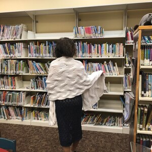 May include: A person wearing a white, textured shawl and a navy blue skirt, standing in a library. The shawl has a wave-like pattern. Bookshelves filled with books are in the background.