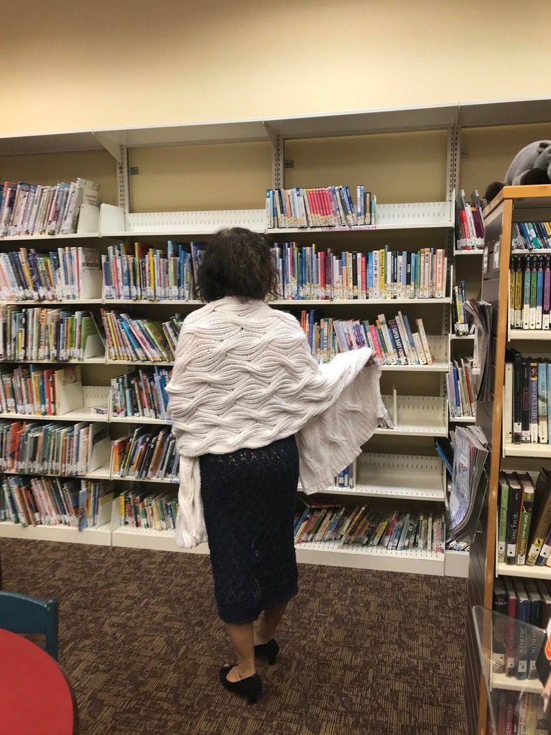 May include: A person wearing a white, textured shawl and a navy blue skirt, standing in a library. The shawl has a wave-like pattern. Bookshelves filled with books are in the background.