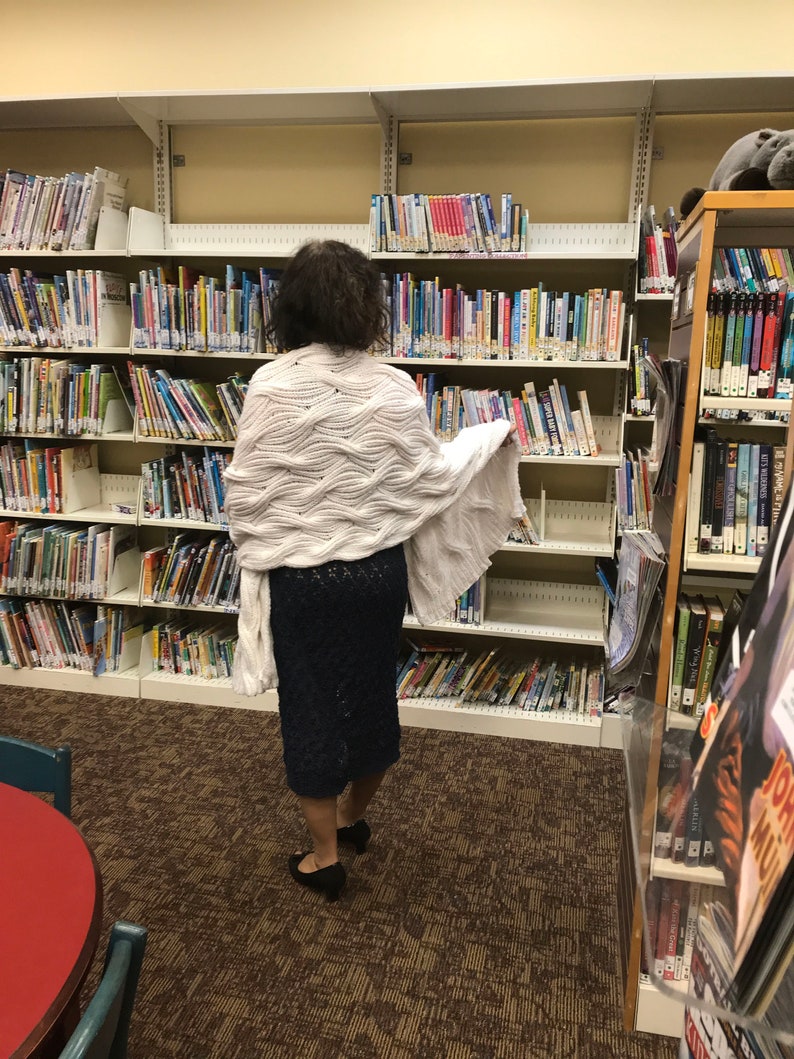 May include: A person wearing a white, textured shawl and a dark blue skirt stands in a library. The library shelves are filled with books of various sizes and colors. The floor is covered with a brown patterned carpet.