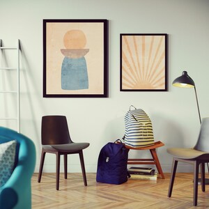 May include: A living room interior with two framed abstract art prints on the wall, a white ladder, two chairs, a blue armchair with a patterned pillow, a floor lamp, and two backpacks on a wooden stool.