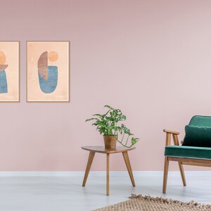 May include: Two framed abstract art prints with geometric shapes in shades of blue, orange, and brown, hanging on a pale pink wall. A green velvet armchair with wooden legs sits in front of a wooden side table with a potted plant.