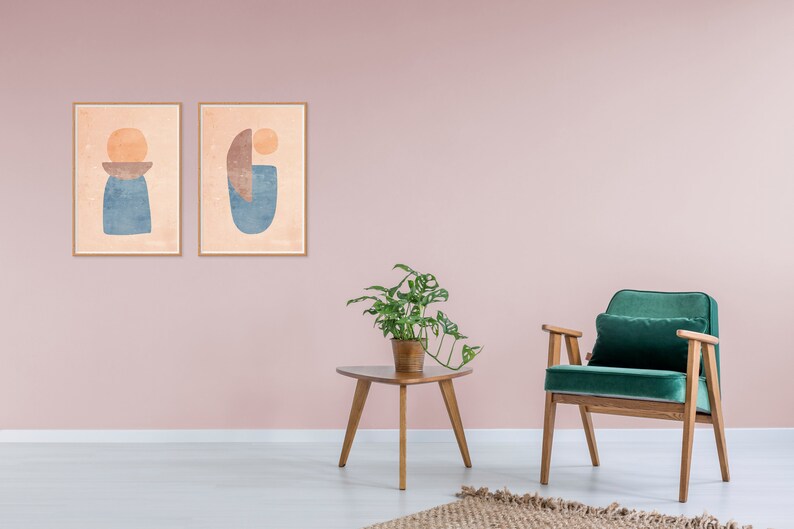 May include: Two framed abstract art prints with geometric shapes in shades of blue, orange, and brown, hanging on a pale pink wall. A green velvet armchair with wooden legs sits in front of a wooden side table with a potted plant.