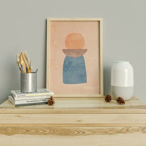 May include: A wooden dresser with two drawers, a white ceramic diffuser, a framed abstract print with geometric shapes in orange, brown, and blue, and a silver metal container with brushes and pencils on top of a stack of books.