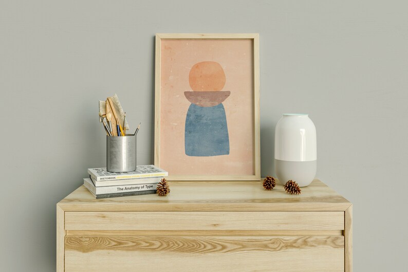 May include: A wooden dresser with two drawers, a white ceramic diffuser, a framed abstract print with geometric shapes in orange, brown, and blue, and a silver metal container with brushes and pencils on top of a stack of books.