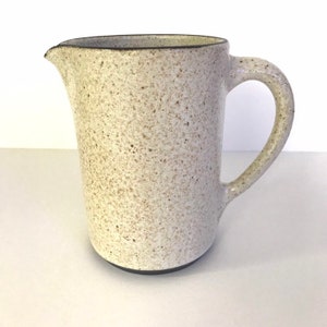 May include: A white ceramic pitcher with a speckled glaze and a black rim. The pitcher has a rounded body and a single handle.