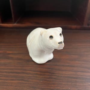 May include: A small, white polar bear figurine with a detailed carved design. The bear has black eyes and a small, dark nose. The figurine is sitting on a dark brown wooden surface.