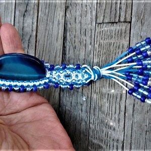 May include: A handmade macrame bracelet with a large, oval, blue gemstone center. The bracelet features a blue and white woven pattern, accented with blue beads. A tassel of blue and white cords with blue and silver beads completes the design.