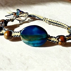 May include: A bracelet featuring a large, oval, blue and green banded stone centerpiece, flanked by brown tiger eye beads and smaller blue beads. The stones are connected by light-colored macrame cord, secured with a silver-toned metal lobster clasp.