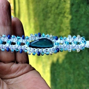 May include: Macramé bracelet with a central teal stone, set within a white macramé design, and adorned with blue and clear beads. The bracelet is displayed against a green backdrop.