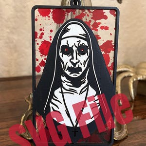 May include: Black and white rectangular air freshener featuring a nun with red eyes and a black habit. The background has red splatter marks. A black cord is attached for hanging. The air freshener is a horror-themed item.