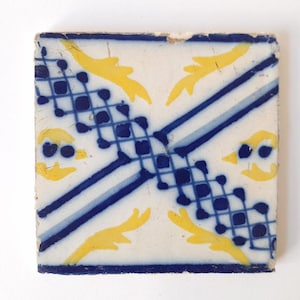 Könnte beinhalten: Eine quadratische Keramikfliese mit einem blauen und weißen geometrischen Muster und gelben floralen Akzenten. Die Fliese hat einen weißen Hintergrund mit einem blauen und weißen Gitterdesign in der Mitte. Die Ecken der Fliese zeigen gelbe florale Motive.