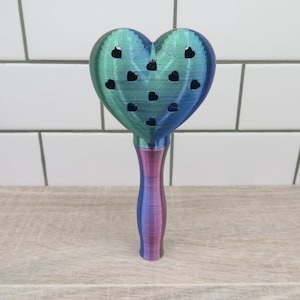 May include: A heart-shaped, teal and purple, 3D printed, decorative object with a handle. The heart has small, black heart-shaped cutouts.