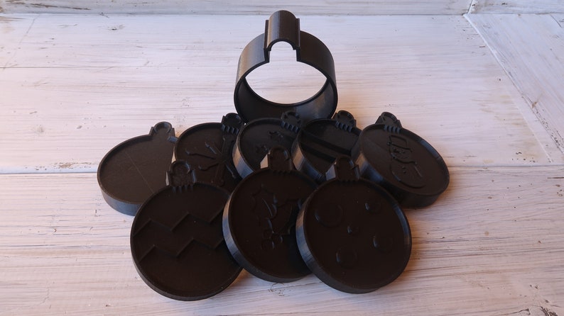 K&ouml;nnte beinhalten: Ein Set aus acht schwarzen Weihnachtsbaumschmuck-Ausstechern aus Kunststoff. Die Ausstecher sind in Form von Ornamenten mit verschiedenen Designs, darunter Schneeflocken, Sterne und ein Schneemann. Ein Ausstecher ist gr&ouml;&szlig;er als die anderen und hat die Form eines traditionellen Weihnachtsbaumanh&auml;ngers.