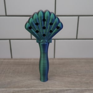 Peut inclure: Un ustensile de cuisine en forme de coquille de couleur verte et bleue avec une longue poignée. L'ustensile est percé de trous dans la coquille et est conçu pour ramasser ou filtrer.