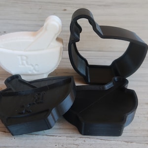 May include: A set of four black and white 3D printed cookie cutters. The cutters are shaped like a mortar and pestle, a baby carriage, a bottle, and a teacup. The cutters are made of a durable plastic and are perfect for making cookies, fondant, or other edible treats.