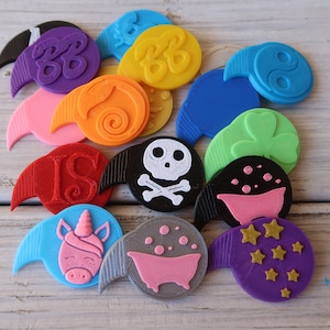 Peut inclure: Une collection de 12 bouchons de bouteille en plastique imprimés en 3D de différentes couleurs avec divers motifs, dont un crâne et des os croisés, une licorne, un trèfle à trois feuilles, une baignoire, un symbole yin yang et des étoiles.