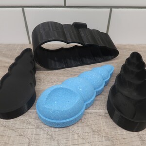 May include: A blue, seashell-shaped bath bomb sits among three black, 3D-printed seashell-shaped molds. The molds are in various sizes and shapes, including a full shell, a half shell, and a ring.