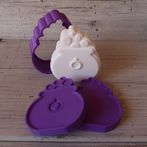 Könnte beinhalten: Drei violette Plastik-Ausstecher in Form eines Goldtopfs. Die Ausstecher liegen auf einer hellbraunen Holzoberfläche.