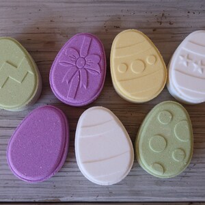 May include: Seven bath bombs shaped like Easter eggs in various colors and designs. The bath bombs are decorated with zigzags, polka dots, stars, and a bow.
