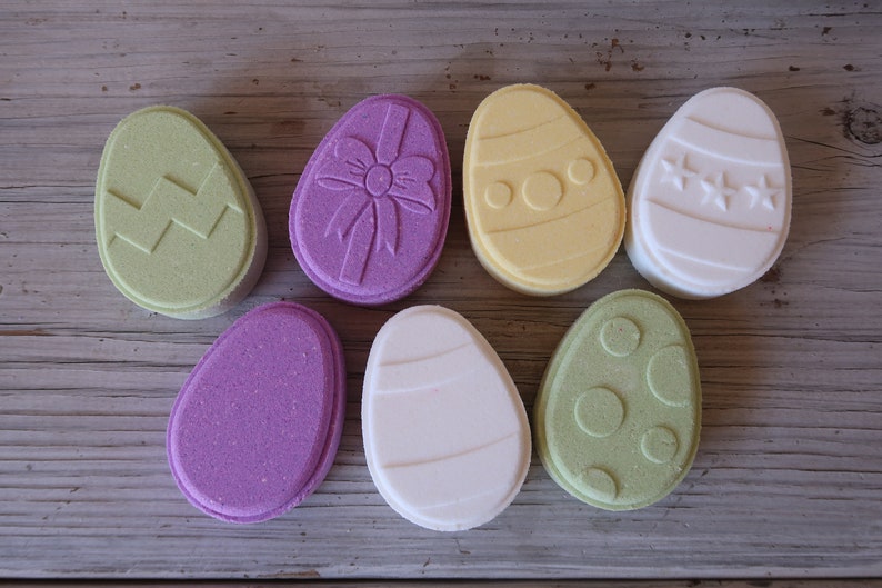 May include: Seven bath bombs shaped like Easter eggs in various colors and designs. The bath bombs are decorated with zigzags, polka dots, stars, and a bow.
