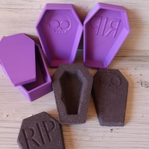 May include: Five purple and brown coffin-shaped molds. Two of the molds are purple and have the words "RIP" and a skull design. Three of the molds are brown and have the words "RIP" and a skull design.