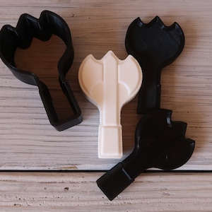May include: Three cookie cutters, two black and one white, shaped like a medieval axe. The white axe is a raised design, while the black cutters are flat.