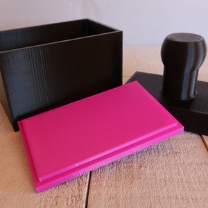 May include: A black rectangular box with a pink lid and a black stamp with a round handle. The box and stamp are made of 3D printed plastic.