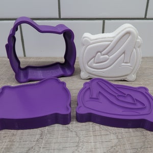 Könnte beinhalten: Ein Set aus lila und weißen Ausstechformen. Das Set enthält eine lila Umrissform, einen lila Abdruck und einen weißen Abdruck eines High-Heel-Schuhs auf einem Kissen. Die Ausstecher liegen auf einer Holzoberfläche.
