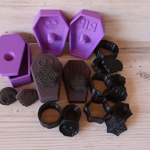 May include: A set of purple and black Halloween-themed cookie cutters. The set includes two coffin-shaped cutters, one with a skull and crossbones design and the other with the word "RIP" on it. There are also several smaller cutters in the shape of skulls, spiders, and webs.