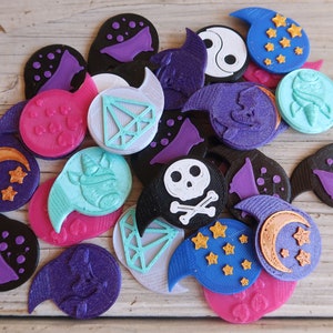 May include: A collection of colorful, 3D printed tokens featuring various designs, including a skull and crossbones, a crescent moon, a diamond, a yin yang symbol, and a unicorn. The tokens are made of plastic and have a smooth, glossy finish.