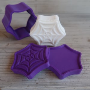 May include: Three purple plastic cookie cutters shaped like a star with a spider web design. One cutter is solid, one has a spider web design, and one is open.