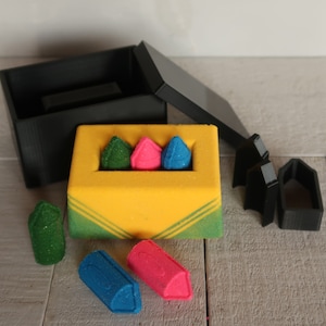 May include: A set of four crayons in a yellow box with green stripes. The crayons are green, pink, blue, and red. The box is open and the crayons are spilling out.