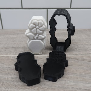 May include: A white plastic flower in a vase cookie cutter with a black plastic outline cookie cutter.