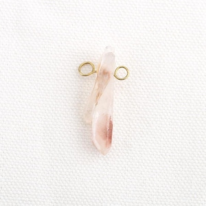 May include: Two clear quartz crystal points with a pinkish hue, each attached to a small gold-colored loop. The crystals are arranged in a vertical orientation against a white background.