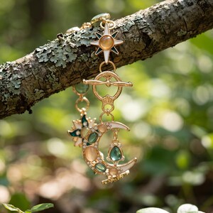 May include: A gold-toned bracelet featuring celestial motifs including a crescent moon and star charms. The design incorporates white enamel, small clear crystals, and oval-shaped cabochon stones in shades of soft peach and translucent green.