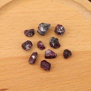 May include: A collection of raw, uncut ruby gemstones. The stones vary in size and shape, displaying a deep red hue with white and gray inclusions. They are arranged on a light brown wooden surface.