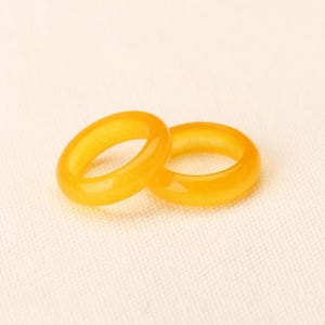 Puede incluir: Dos anillos translúcidos de color ámbar se muestran sobre una superficie color crema. Los anillos son lisos y pulidos, con forma redondeada. Los anillos están hechos de un material de piedra preciosa y están colocados cerca uno del otro.