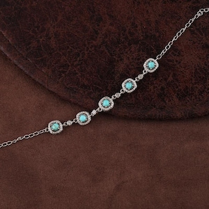 Pode incluir: Uma pulseira de prata com cinco pedras turquesas quadradas, cada uma rodeada por pequenas pedras preciosas transparentes. A pulseira &eacute; exibida em uma superf&iacute;cie marrom, destacando sua delicada corrente e design intrincado.